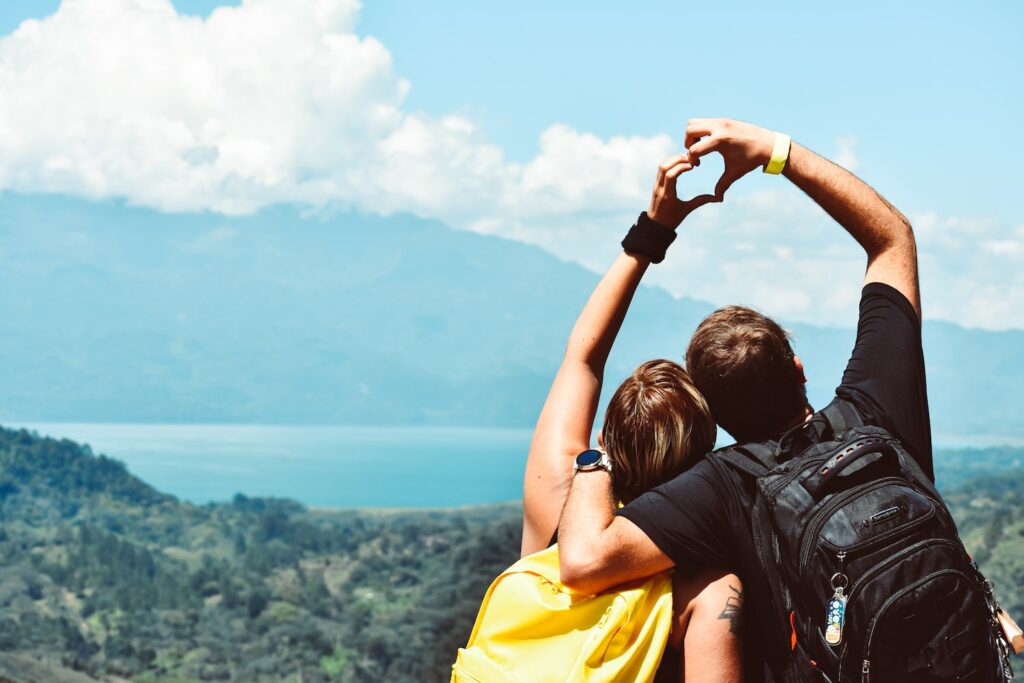 A couple make a heart sign with their hands whilst looking out at a beautiful landscape